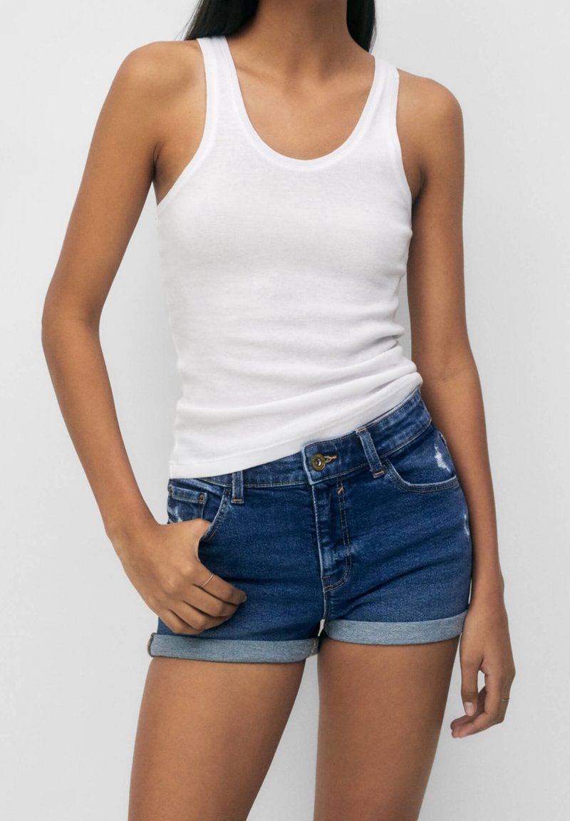 Vit bomullstanktopp i kombination med blå denimshorts. Shortsen har en upprullad fåll och framficka, med lätt slitage på tyget.