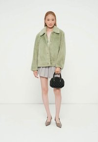 Veste en fausse fourrure verte avec col et fermeture à boutons ; portée sur un top clair et une jupe plissée. Le mannequin tient un sac à main noir côtelé.