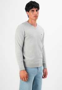 Férfi világosszürke V-nyakú pulóvert és világoskék farmert visel, egy fehér háttér előtt áll, enyhén oldalra nézve.