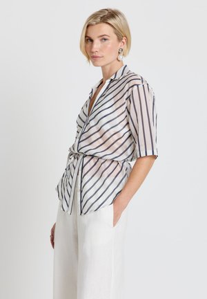 Blonde woman wearing oversized striped blouse with three-quarter sleeves and white high-waist pants, posing with one hand in pocket.