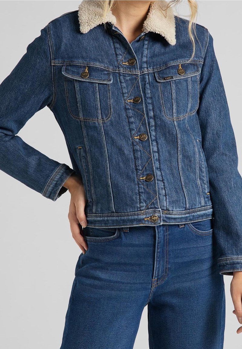 Femme portant une veste en denim bleue avec col en polaire et un jean bleu assorti, main posée sur la hanche.