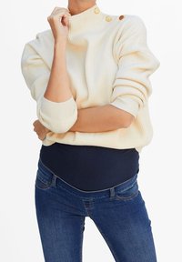 Suéter crema de canalé con cuello alto y tres botones dorados, combinado con jeans de maternidad azul oscuro que tienen bolsillos frontales.
