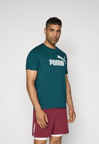 Teal kortærmet T-shirt med et stort hvidt "PUMA" logo, parret med bordeaux atletikshorts med hvide sideakcenter.