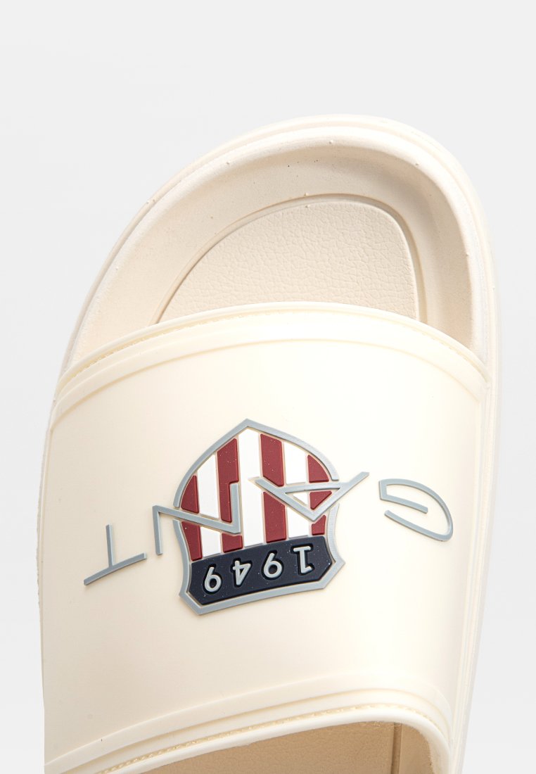 Cremfarbene Rutschsandale mit einem strukturierten Riemen, grauem Logo mit der Aufschrift "GANT" und einem Wappenemblem mit roten und weißen Streifen und "1949".