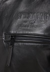 Black leather material with embossed "MUSTANG JEANS COMPANY" logo. Features a zippered pocket with a textured pull tab and stitched edges.