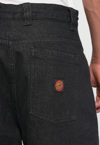 Calças de ganga pretas com um patch centrado em vermelho e amarelo "Santa Cruz" no bolso de trás. O tecido tem um acabamento texturizado.