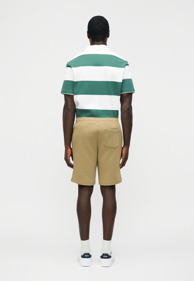 Άνδρας που στέκεται με την πλάτη, φοράει πράσινη και λευκή ριγωτή κοντομάνικη μπλούζα, μπεζ σορτς με πίσω τσέπη, λευκές κάλτσες και λευκά αθλητικά παπούτσια.