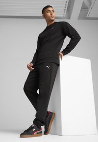 Svart Puma-hoodie med en liten logotyp, matchande svarta byxor och svarta sneakers med röda och vita detaljer. Slät tygkvalitet, avslappnad passform.