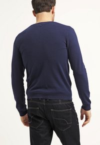 Marinblå tröja i stickad kvalitet, med rundad halsringning, långa ärmar och ribbade ärmhål. Bärs med mörka jeans, sedd bakifrån.