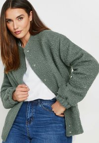 Olive green, fluffy fleece jacket with a rounded neckline, snap buttons, and elasticized cuffs, worn over a white shirt and blue jeans.