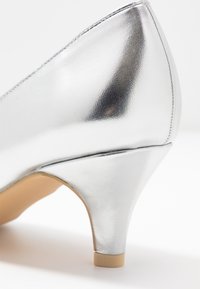 Escarpins argentés avec une finition métallique brillante, bout pointu, et un talon épais de hauteur intermédiaire. Texture lisse et détails de couture soignés.