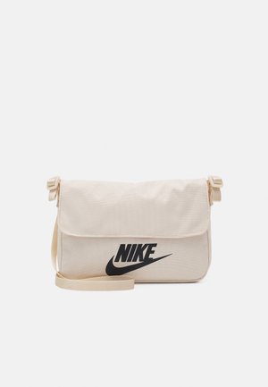 Beżowa torba na ramię Nike z regulowanym paskiem i zamknięciem na klapkę, z dużym czarnym logo Nike i znakiem swoosh z przodu.