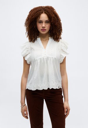 Junge Frau mit lockigem Haar, die eine weiße Lochstickerei-Bluse mit Rüschenärmeln und braune Hose trägt, vor einem schlichten Hintergrund stehend.