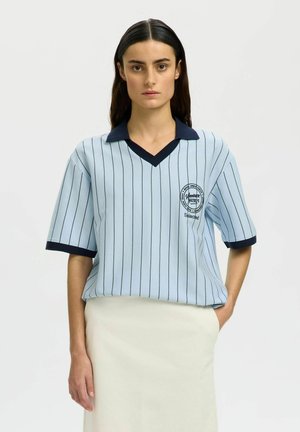 Femme aux longs cheveux foncés portant une chemise à manches courtes bleu clair à fines rayures avec col et poignets foncés, associée à une jupe crème, debout face à l'appareil.