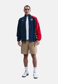 Nike Jacke in Marineblau und Rot, mit Reißverschluss und Logo. Getragen mit beigen Cargo-Shorts, T-Shirt in Weiß und weißen Sneakers mit schwarzen Akzenten.