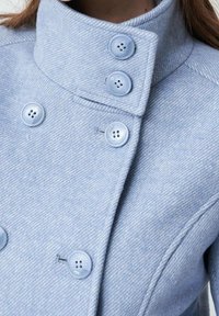 Cappotto in lana azzurro chiaro con colletto alto e sei bottoni abbinati, indossato da una persona con i capelli castani visibili sul bordo superiore.