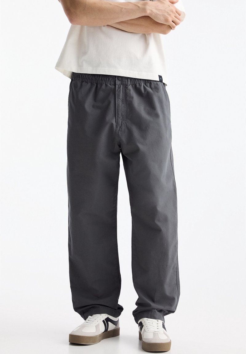 Personne portant un pantalon ample gris foncé, des baskets blanches avec des bandes noires, debout les bras croisés sur un fond blanc uni.