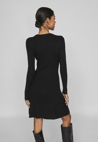 Μαύρο πλεκτό φόρεμα με ραβδώσεις, μακριά μανίκια, εφαρμοστό μπούστο και φουσκωτό μακρύ φούστα. Λείος υφή με μήκος στο μέσο του μηρού και διακριτικά πλαϊνά σκισίματα.