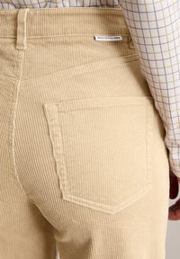 Beżowe spodnie ze sztruksu z pionowym prążkowaniem, z tylną kieszenią i metką marki, zestawione z bluzką w kratę.