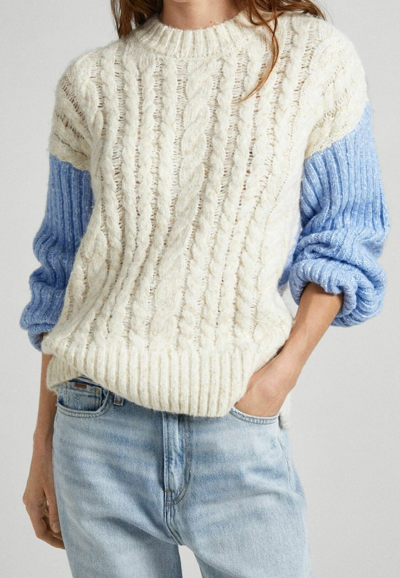 Femme portant un pull à torsades crème avec des manches côtelées bleu clair et un jean bleu clair, une main dans la poche.
