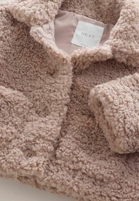 Miękka różowa teddy płaszcz z kręconym, teksturowanym materiałem, zapięcie na guziki i widoczną etykietą marki "NEXT" wewnątrz kołnierza.