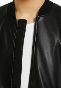 Giacca bomber nera in pelle con una texture liscia, colletto a costine e zip frontale. Indossata su una maglietta bianca, che mette in evidenza un design minimalista.