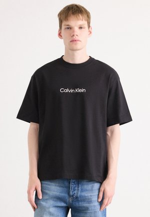 T-shirt noir en coton à manches courtes arborant un logo "Calvin Klein" blanc sur le devant, un col rond et une coupe décontractée. Porté avec un jean bleu.