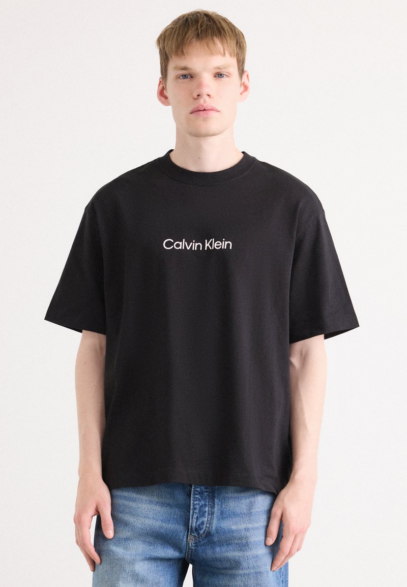 Svart kortermet t-skjorte i bomull med hvit "Calvin Klein" logo på framsiden, rund hals og løs passform. Brukes sammen med blå jeans.