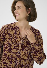 Chemise à manches longues avec un motif de feuilles bordeaux et beige, fermeture à boutons et poignets élastiqués. Matériau léger et coupe décontractée.