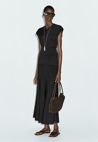 Top nero aderente a maniche corte abbinato a una gonna nera fluente. Gli accessori includono occhiali da sole scuri, una lunga collana d'argento e una borsa in suede marrone.
