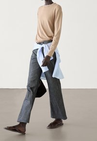 Personne marchant portant un haut beige à manches longues, un pantalon gris à jambes larges, une chemise blanche nouée autour de la taille, et des sandales marron.