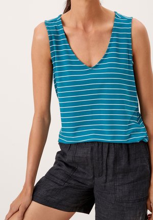 Kvinde iført ærmeløs teal top med hvide vandrette striber og mørkegrå shorts, stående med den ene hånd hvilende på låret.