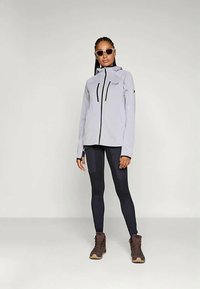 Ženska v svetlo sivi jakni z zadrgo, črnih leginsih, rjavih pohodniških čevljih in belih sončnih očalih stoji ob enotni svetli postavitvi.