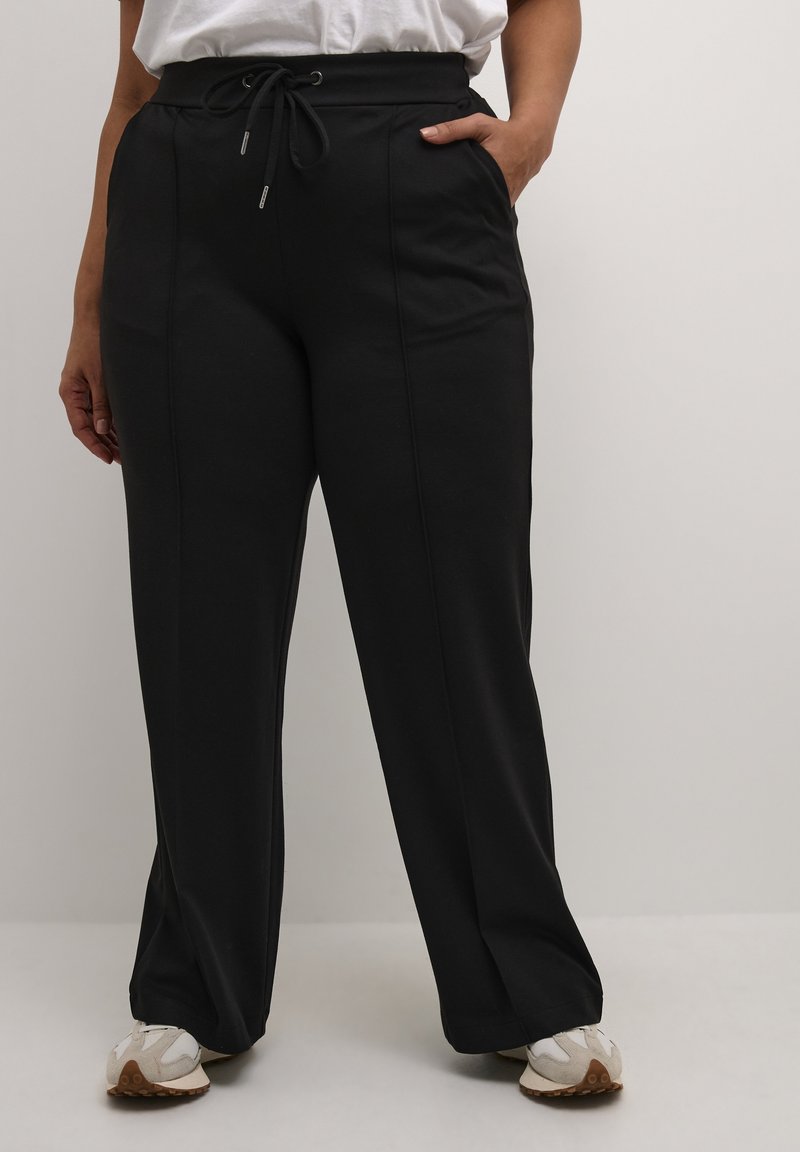 Personne portant un pantalon large noir à taille haute avec un cordon, des poches latérales et des baskets blanches, debout devant un fond clair uni.