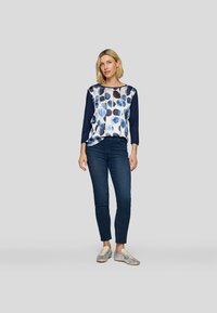 Blaue und weiße gemusterte Bluse mit Dreiviertelärmeln, kombiniert mit dunklen Jeans und silbernen Turnschuhen. Glattes Material mit kreisförmigen Mustern.