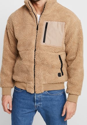 Mężczyzna w beżowej kurtce sherpa fleece z wysokim kołnierzem, beżową kieszenią na piersi na zamek i niebieskich dżinsach.