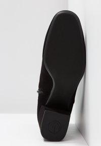 Chaussure en daim noir vue de la semelle, avec un bout carré et un petit talon carré, positionnée verticalement contre un mur blanc d'angle.