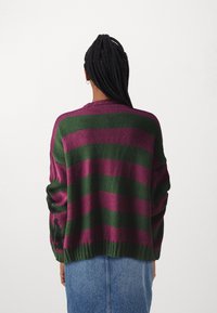 Strickjacke mit kräftigen lila und grünen Streifen, lässiger Schnitt, gerippter Saum und lockere Ärmel. Die Textur wirkt weich und gemütlich.