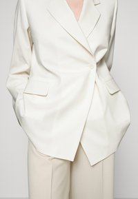 Biała dopasowana marynarka na jeden guzik, z klapowym kołnierzem i bocznymi kieszeniami, zestawiona z beżowymi spodniami. Gładka tekstura tkaniny.