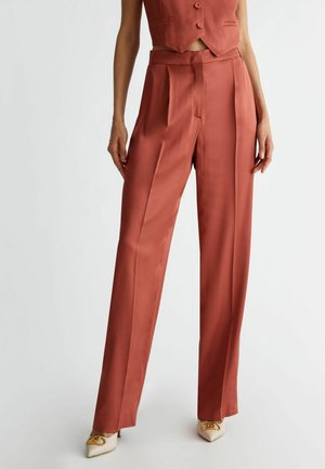 Femme portant un pantalon large taille haute couleur rouille et un gilet court assorti, assortis à des talons blancs pointus avec une boucle dorée.