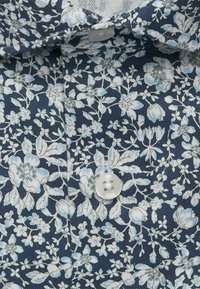 Tissu bleu avec un motif floral dense en blanc et bleu clair, présentant un gros plan d'un bouton blanc à quatre trous et du bord du col.