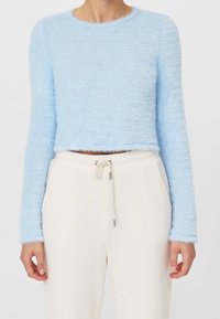 Mujer vestida con un top corto de manga larga fuzzy azul claro y pantalones de chándal crema con cordón, de pie frente a un fondo liso.