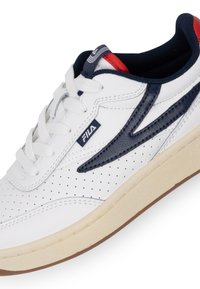 Sneaker atletica bianca con accenti blu navy, dettagli perforati, chiusura con lacci e suola in gomma testurizzata. Logo Fila sul lato.