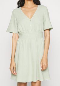 Světle zelené šaty vyrobené z měkké, texturované látky. Mají výstřih do V, krátké rukávy, knoflíkovou légu a přiléhavý pas se širokou sukní.