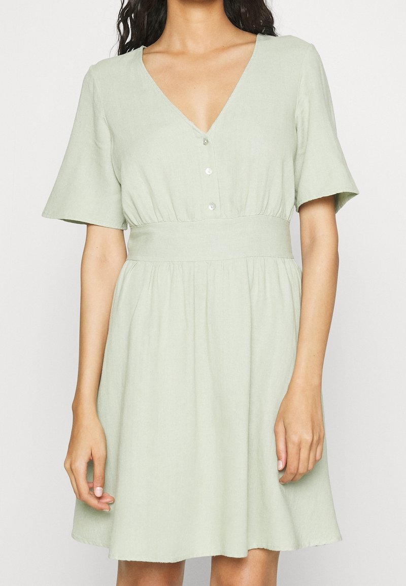 Světle zelené šaty vyrobené z měkké, texturované látky. Mají výstřih do V, krátké rukávy, knoflíkovou légu a přiléhavý pas se širokou sukní.