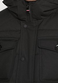 Schwarze Pufferjacke mit hohem Kragen, Reißverschluss, zwei vorderen Klapptaschen und schwarzen Druckknöpfen; glatte, matte Textur.