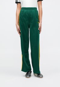 Zielone spodnie sportowe wykonane z gładkiej tkaniny, z żółtymi paskami po bokach i elastycznym pasem. Do tego czarne buty piłkarskie.