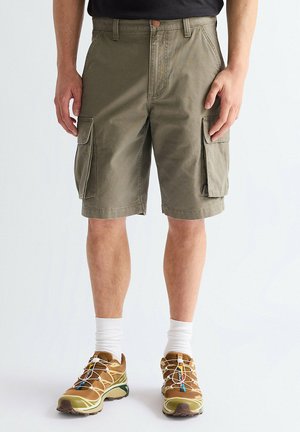 Mann trägt olivgrüne Cargo-Shorts, weiße Sportsocken und braune Trailrunning-Schuhe, steht vor einem schlichten Hintergrund.