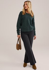 Pull en maille vert foncé avec un motif en losanges texturé, associé à un pantalon large noir et des bottines en daim marron, portant un sac marron.