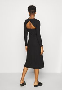 Svart ribbade långärmad klänning med längd till mitten av vaden och en skuren baksida. Slät textur och åtsittande siluett. Bärs med svarta sandaler.